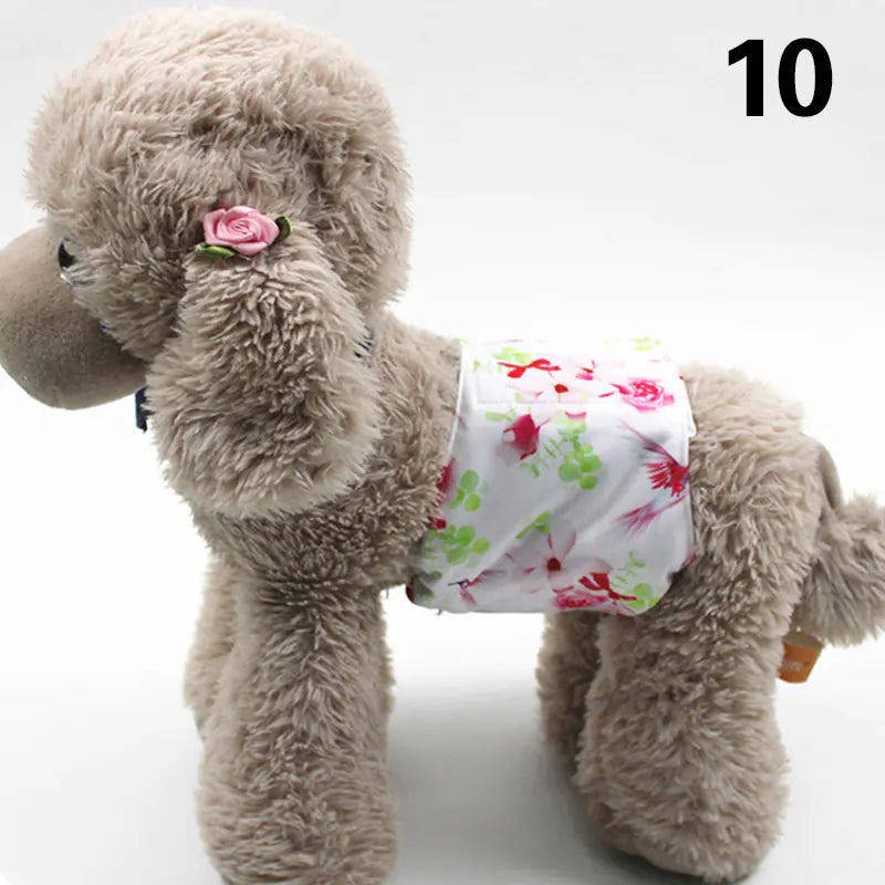 Plush gray poodle dog with a pink flower on its head, wearing a floral printed diaper or pad, positioned in a studio setting with a plain white background.
